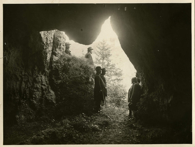 The picture shows six people in the Klausen caves. It is a black and white photograph, probably from the first half of the 20th century. The people are dressed in old, ankle-length clothes and wear caps and hats. The group is standing in the entrance area of the cave and is surrounded by rock walls. You can see the uneven walls and ceilings formed by natural forces.